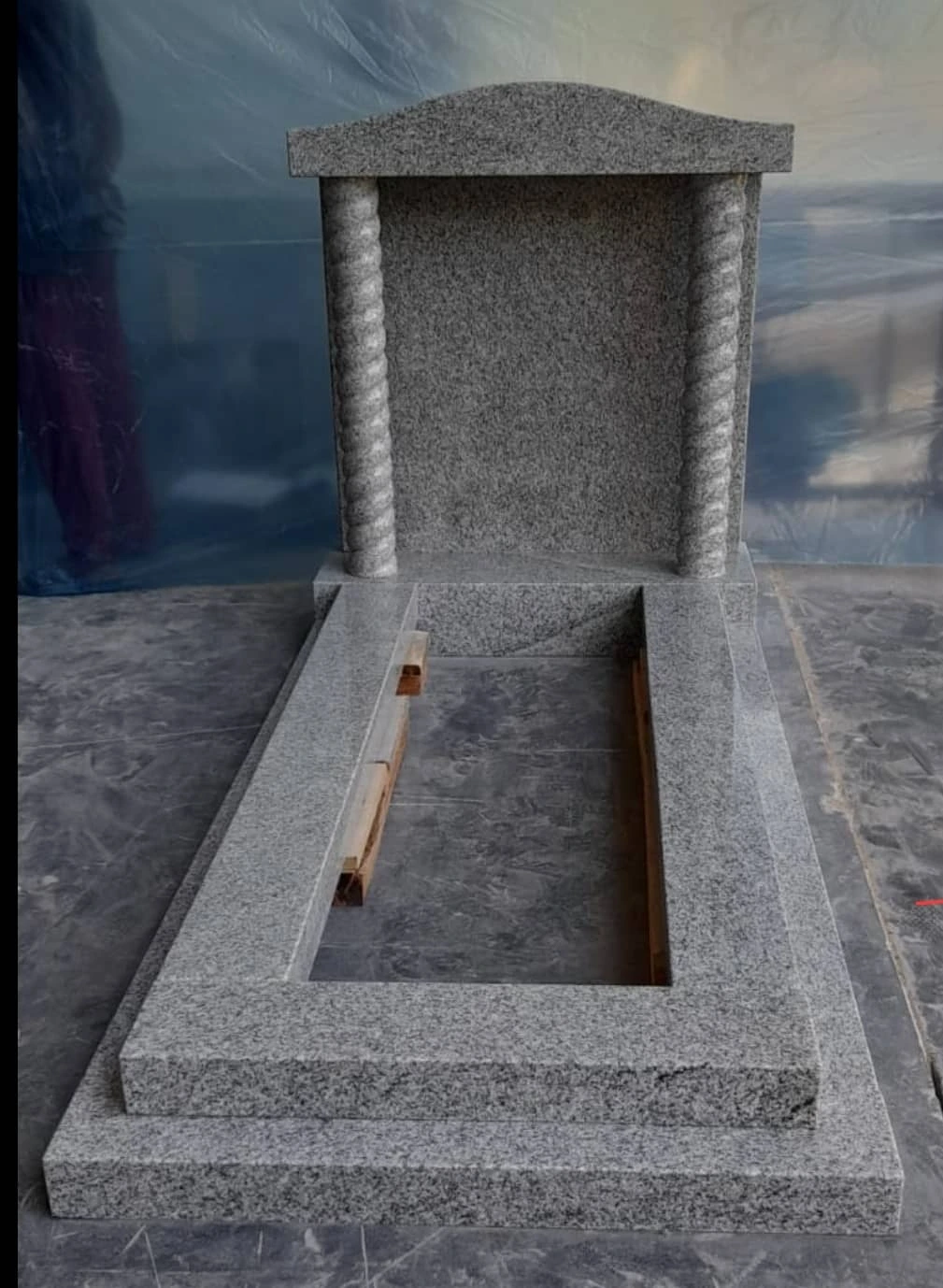 Grey memorial with a capped headstone, column details and matching kerb surround, shown in the workshop